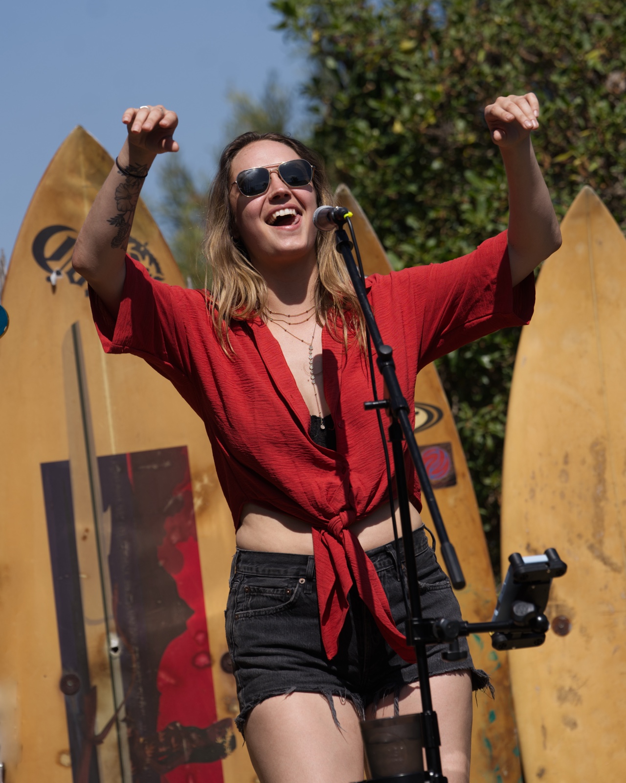 Singer cheering with raised arms in front of a wall of surfboards at an outdoor daytime show