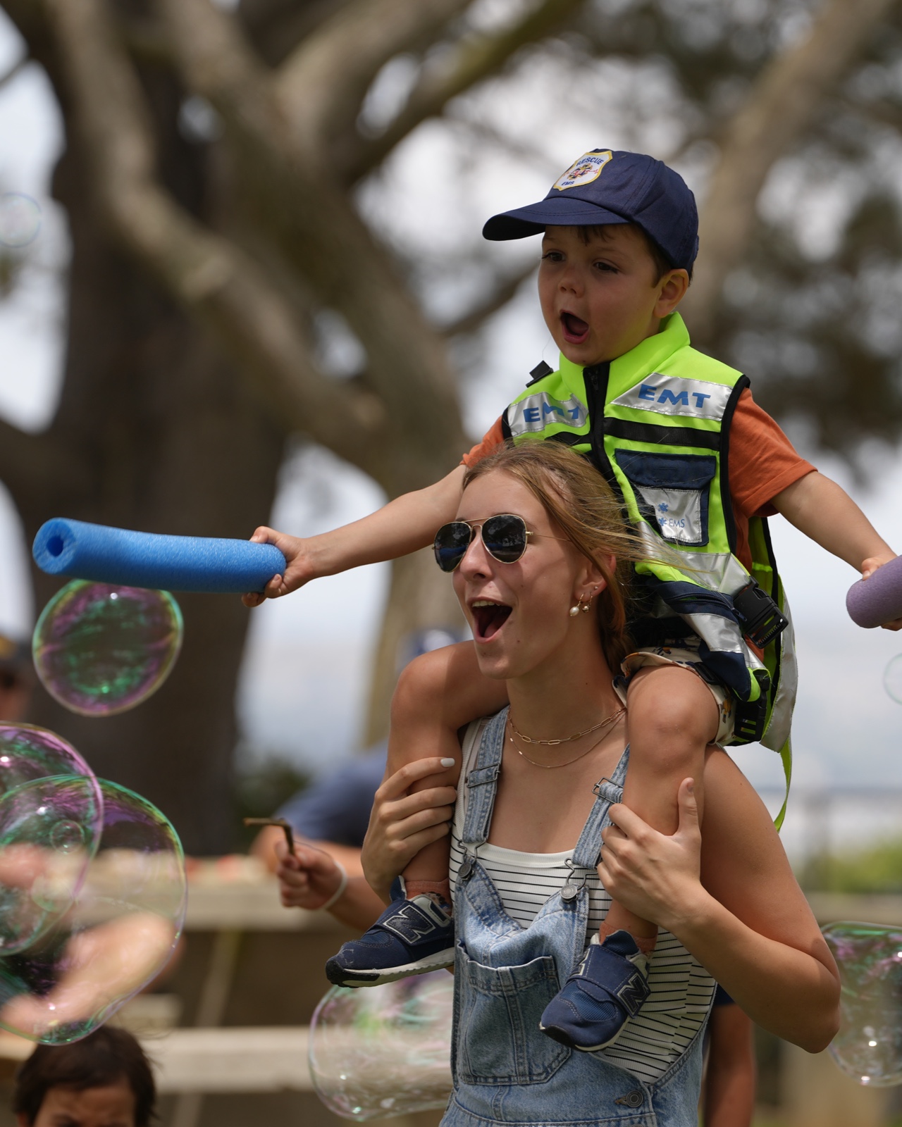 Toddler in a play EMT vest riding on his mom's shoulders while she laughs, blowing bubbles at a park